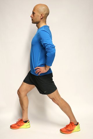 A man standing with his right leg, slightly bent, in front of him. His left leg is stretched out straight behind him, with the heel flat on the floor